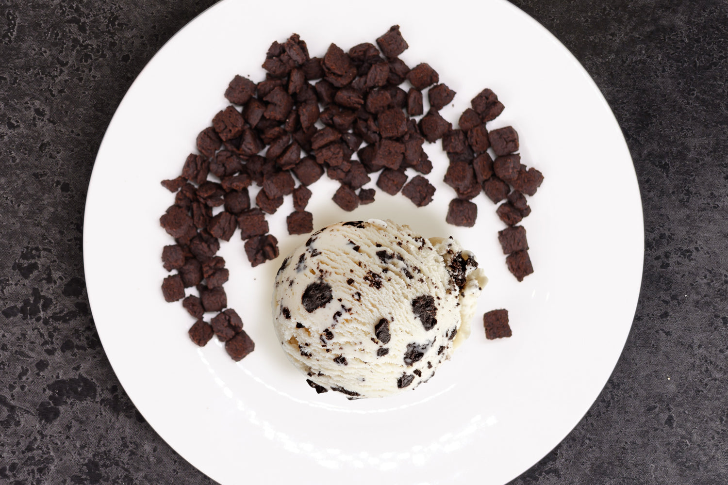 Ice cream scoop with chocolate brownie pieces on a white plate against a dark background. Dessert Kitchen Inclusions